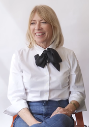 Kim Gordon, smiling and wearing a white blouse with black ribbon and jeans.