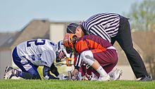 Two men wearing helmets, short-sleeved jerseys, shorts and gloves, one in a maroon and orange color scheme and the other in blue and white, kneel and crouch on grass as a man wearing a black and white vertically-striped long-sleeved shirt and long black pants with a black cap leans from behind them and places a small ball between the mesh heads of two sticks they hold on the ground between them.