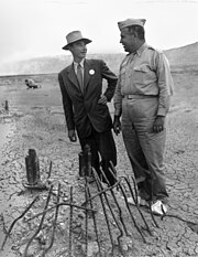 Two men, one in a suit and hat and the other in military uniform, stand in front of twisted metal whilst wearing white overshoes