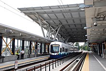 A train station with bare platforms and a tiered roof. A train is seen coming towards the camera on the opposite platform.