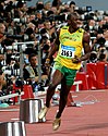 Jamaican athlete Usain Bolt wearing a yellow running vest and green running shorts, with his tongue out, celebrating as he slows down after winning the 2008 Olympic Games 100m final in a record-breaking time