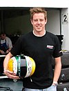 A man in his early twenties standing in a pit lane and smiling at the camera. He is holding a crash helmet in a yellow, dark green and white colour scheme in his right arm.
