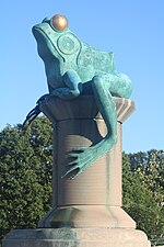 A green copper frog with gold eyes perched atop a giant concrete spool