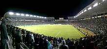 A stadium with a soccer field surrounded by a full crowd. The field is lit with large floodlights above the stands.