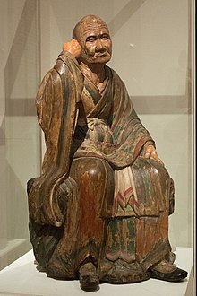 Monk with stern face and slight protrusion in skull, sitting and holding his hand as though he is holding a staff