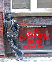 A statue depicting a young Lennon outside a brick building. Next to the statue are three windows, with two side-by-side above the lower, which bears signage advertising the Cavern pub.