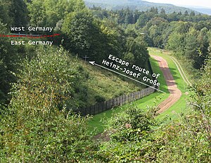 A lush, green, wooded landscape through which the border line about 10 metres (32 ft) wide, cleared and grassed with a narrow dirt strip in the middle, runs from bottom-left across the image, turning slightly to run directly away from the viewer into the distance. To the right is the outer fence and a steep incline leading up to the near-side fence of a West German road. A schematic arrow, marked "Escape route of Heinz-Josef Großze", points to a memorial cross at the point of the shooting, half way up the incline.