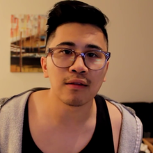 Tong, wearing his glasses and a Sleeveless shirt underneath a hoodie jacket, is looking straight at the camera from his living room.