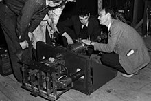 Four men in suits bend over a piece of machinery.