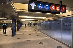A mezzanine within the 59th Street–Columbus Circle station. There are storefronts to the right. A sign with an arrow, "A", "C", "B", "D", and "1" icons hangs from the ceiling.