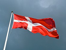 A flag flies from a flagpole against the sky. The flag is red, divided in four by a white cross, the vertical bar of which is shifted toward the flagpole.