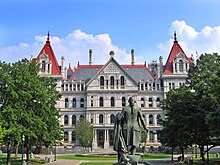 A metallic statue of a man with a short ponytail in his hair, seen from behind, wearing a long coat and holding clothes in his left arm and a walking stick in his right. He is facing a tall stone building in the distance with red peaked roofs. On either side of the image is a row of trees