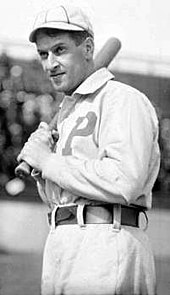 A man in a white baseball uniform and white baseball cap holds a baseball bat against his right shoulder while looking to the left of the image. His uniform has a large block "P" on the chest, and he has a slight smirk on his face.