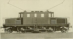 Side view of an electric locomotive with prominent coupling rods between its three driving wheels and a triangular coupling rod from the two motors. There are single-axle pony trucks at each end.