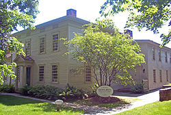 A brown wooden house with wing, lit by the sun from above. At the foot of a small tree in front of the building at the center, an oval sign identifies it as the Tapping Reeve House and Law School