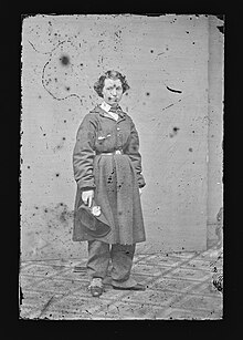 A young woman stands facing the camera in front of a plain backdrop. Her complexion is white, and her short bobbed hair is parted and combed to her left, with curls ending above her ears. She wears an overcoat, belted at the waist, that ends at the knee, over matching pants. She holds a hat in her right hand, and is wearing spats over her closed flat leather shoes. This image is in black and white, and there are marks of deterioration, including scratches, spots and small losses at the edges.