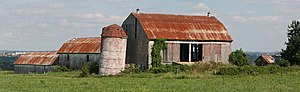 A short silo in the centre of the photograph is slanted slightly to the right, topped by a conical red roof. Three barns form a V shape behind the silo. To its right is a large barn, with slanted red roof and open doors. Perpendicular to it are two similar but smaller barns in series, visible to the left of the silo.