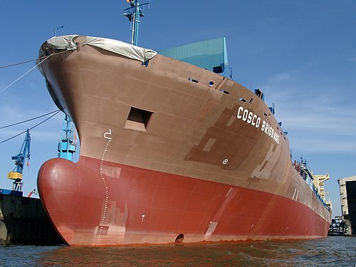 A photograph of the rusty brown hull of a container ship, docked in a shipyard.