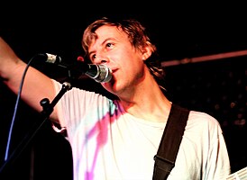 John Vanderslice wearing a white t-shirt, standing onstage, holding an electric guitar, and singing into a microphone