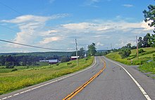 A two-lane highway in a sparsely developed and hilly area with two farm buildings nearby. Large mountain ridges are visible in the distance.