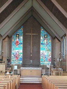 The interior of a building, with a vaulted ceiling supported by wood beams. The far end shows the back wall, which has a large wooden cross hung on a wall between two stained glass windows. An altar is visible before the wall, with a central path leading from it to the foreground, adjacent to which are rows of pews.