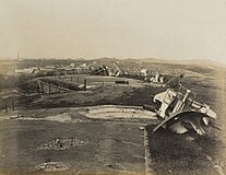 青岛战役后的台西镇炮台，照片自西掩蔽部向东拍摄，可见3门210毫米加农炮残骸以及中央的炮兵观测台，后面远处可见台西镇与市区各山丘