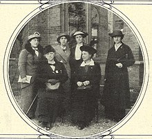 Six women in long winter coats wearing hats standing in front of a building