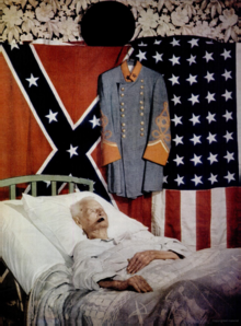 An old man resting in a bed below a Confederate flag, a gray confederate uniform, and a 48-star American flag