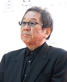 Takahashi in three-quarter profile at press event wearing black suit and black shirt and glasses