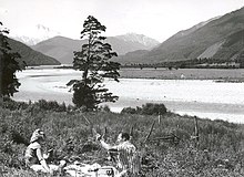 a couple having a picnic on a grassy slope overlooking river flats and snowy mountains;the woman has sunglasses, the man a striped shirt and a fishing rod. A transistor radio sits between them.