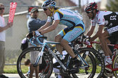 Two cyclists, one wearing a blue and white jersey with yellow trim and the other, to his right, a black and white jersey with red trim, race down a road while spectators watch from beside them