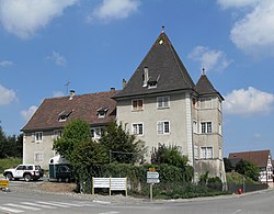 A grey building, in two sections, one two storeys tall with tall, angled, brown tiled roof, the other two intersecting square towers, three storeys tall, each with tall square-pyramidal slate roofs.