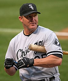 A man wearing a gray baseball uniform with white and black trim and a black baseball cap with "Sox" in white Old English lettering on the face holding a baseball bat under his left arm