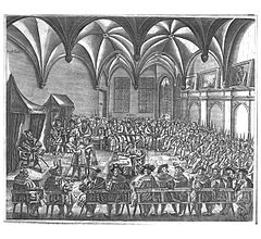 Men gather in a large room, seated on benches around an open center space. Two men read a document to another man seated on a throne.