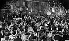 Large, ornate room, filled with people in formal dress sitting at different tables.