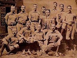 A sepia photograph of twelve men arranged in two rows, standing and sitting. Ten are wearing light baseball uniforms with dark socks, while two are dressed in suits.