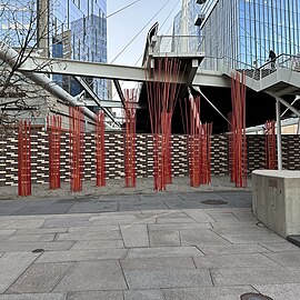 An outdoor urban installation consisting of multiple thin red rods arranged in clusters, creating a striking visual contrast against the gray paving stones, a brick wall, and the metal framework of an overhead pedestrian bridge.