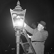 A lamplighter climbs a ladder to light a gas streetlight.