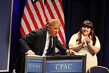 Trump, leaning heavily onto a lectern, with his mouth open mid-speech and a woman clapping politely next to him