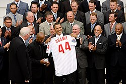 Several men of varying ages wearing suits clap as a man in the center of the photo smiles holding up a white baseball jersey which reads "OBAMA 44" on the back.
