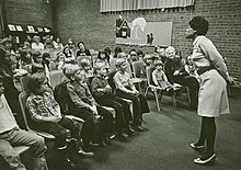 Rita Cox standing and telling story in front of room of seated children
