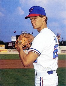 A man wearing a white baseball jersey and a blue cap with a white "N" on the center stands to one side with his hands bought together in his glove.