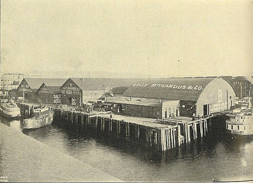 Published in 1900 in the book Seattle and the Orient, this shows Pier B (presumably still the "old" Pier B, because the shed structure matches) in use by the Lilly Bogardus agricultural company. There is a large "B" on the end of the pier shed and smaller letters under that not entirely legible beyond "Lilly Bo…" but consistent with the inscription in the following photo. Between that pier shed and the shore are three sheds at right angles to that, numbered (left to right) "1", "3", and "2", all associated with Lilly Bogardus.