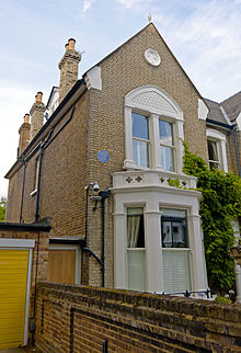 A two-story beige brick house with pointed roof topped by several chimneys with pots and a wooden bay window on the front at ground level. In front is a brick wall. There is a circular blue plaque on the left side of the house near the second storey