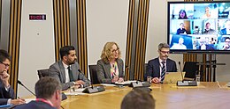 In a Scottish Parliament Committee room, Slater and Yousaf meet sit at a large table of business representatives, with screens flanking them with virtual attendees.
