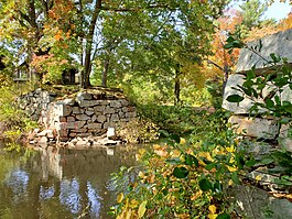 Stone bridge abutments on both sides of a small river with trees with autumn colors mostly hiding a wooden house