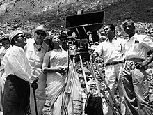 Black-and-white image of cast and crew outdoors around a film camera