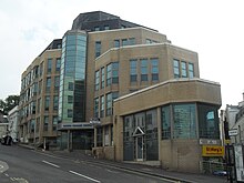 A building, with a modern look in design, stands on a hilly corner between two streets. Each layer of the building juts out below the one above it. The bricks are of a tan color, and a sign says, "Queen Square House", under a portion of glass in the building.