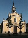 A church with a yellow facade and white architectural details. It features a central wooden door flanked by columns and two small trees. Above the door is a large arched window, and the church is topped with a bell tower with a cross at its peak. The building is situated on a street with a clear blue sky in the background.