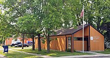 Small one story wooden building surrounded by trees.